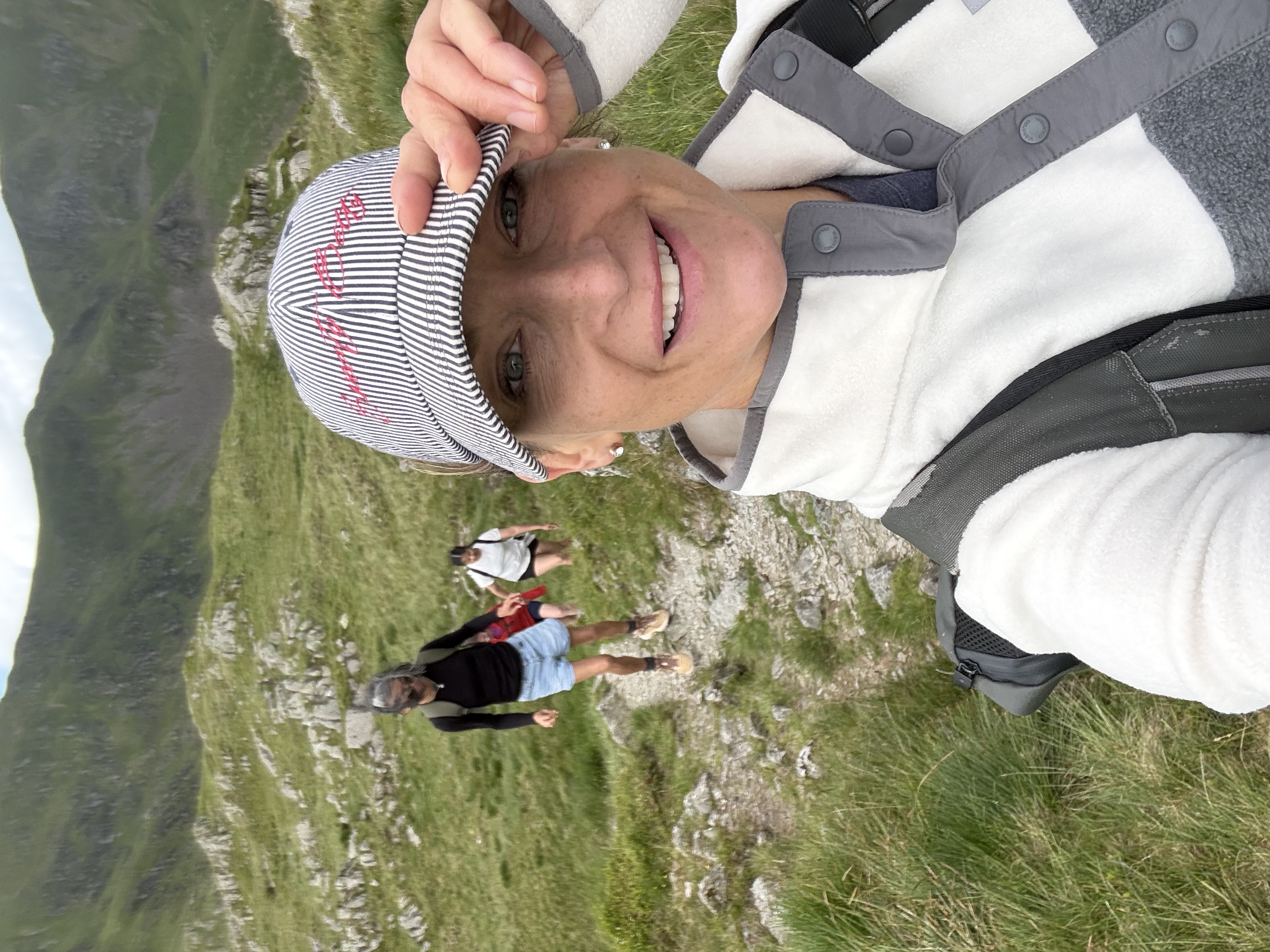 Selfie of a woman in a striped cap with the group climbing the fell behind her