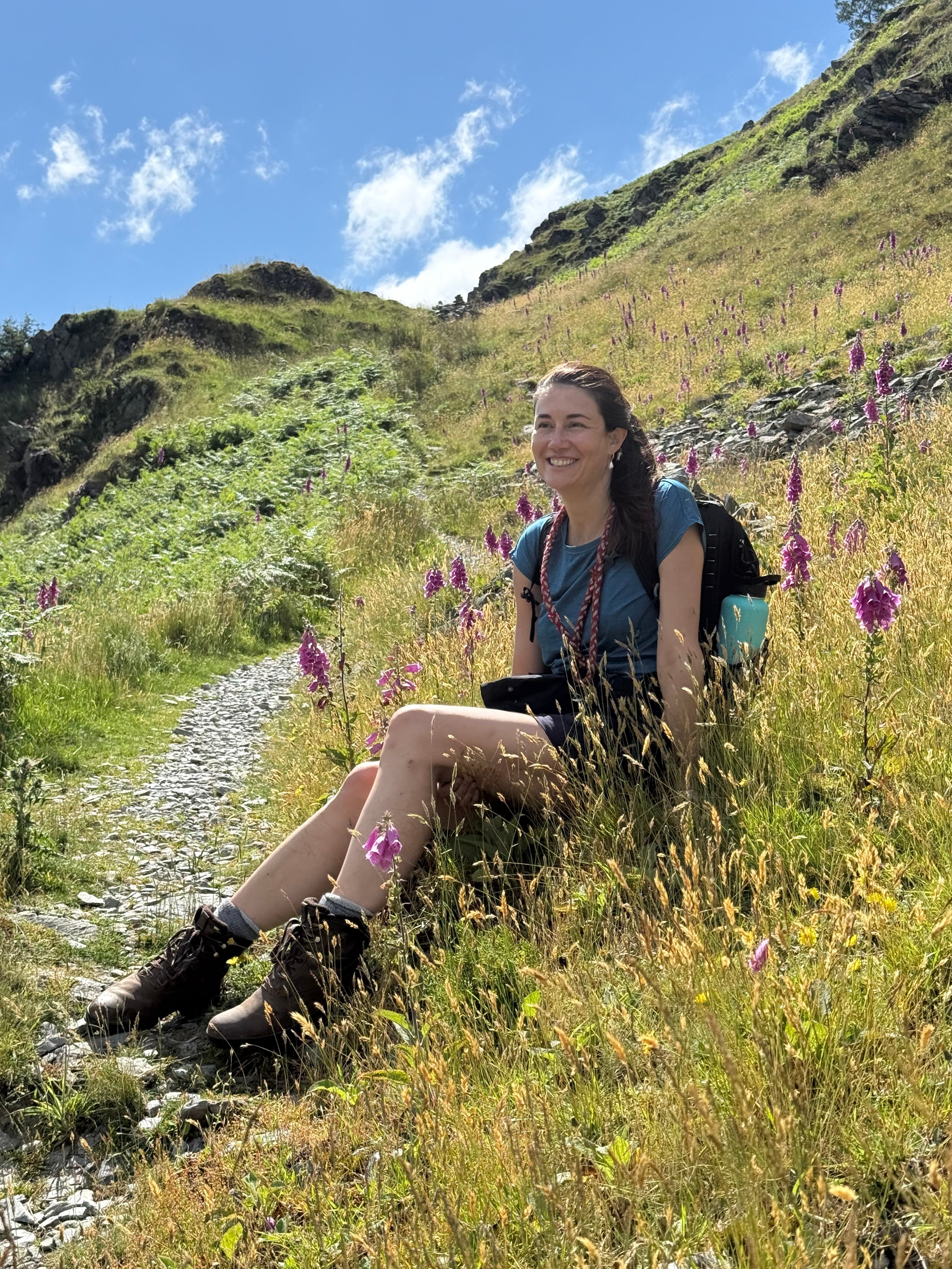 Smiling woman with a backpack sitting among purple foxgloves and golden grasses beside a mountain path