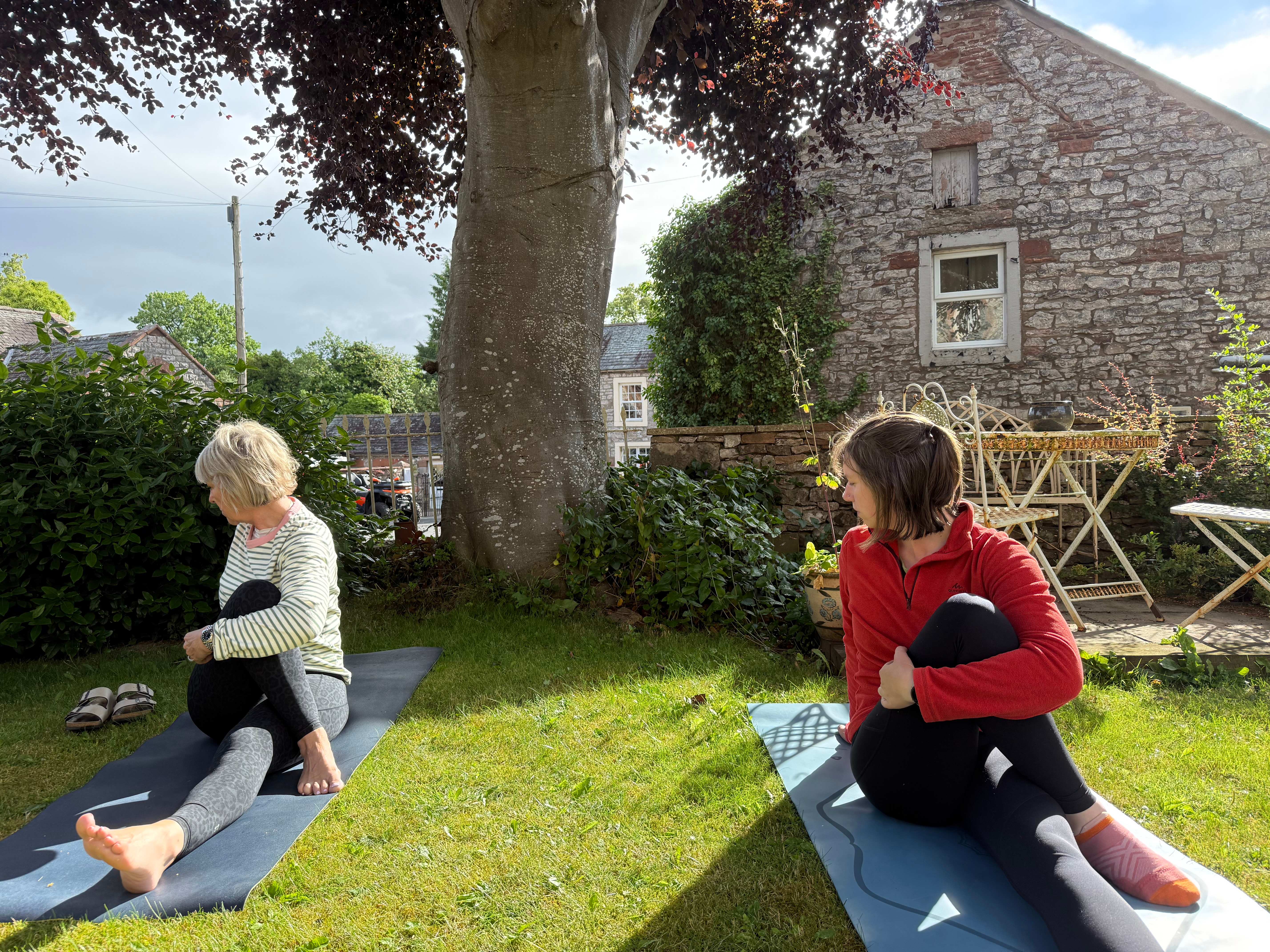 Two women in a seated yoga pose on mats in a sunny garden beside a stone farmhouse