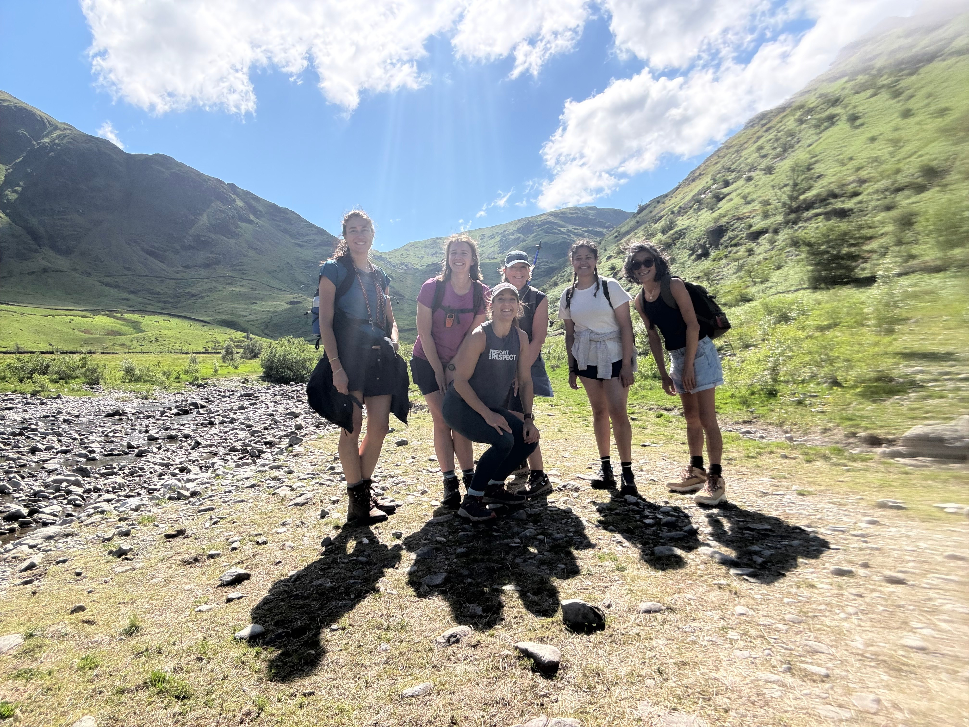Group of six women posing together on a rocky riverbed in a green valley under blue skies