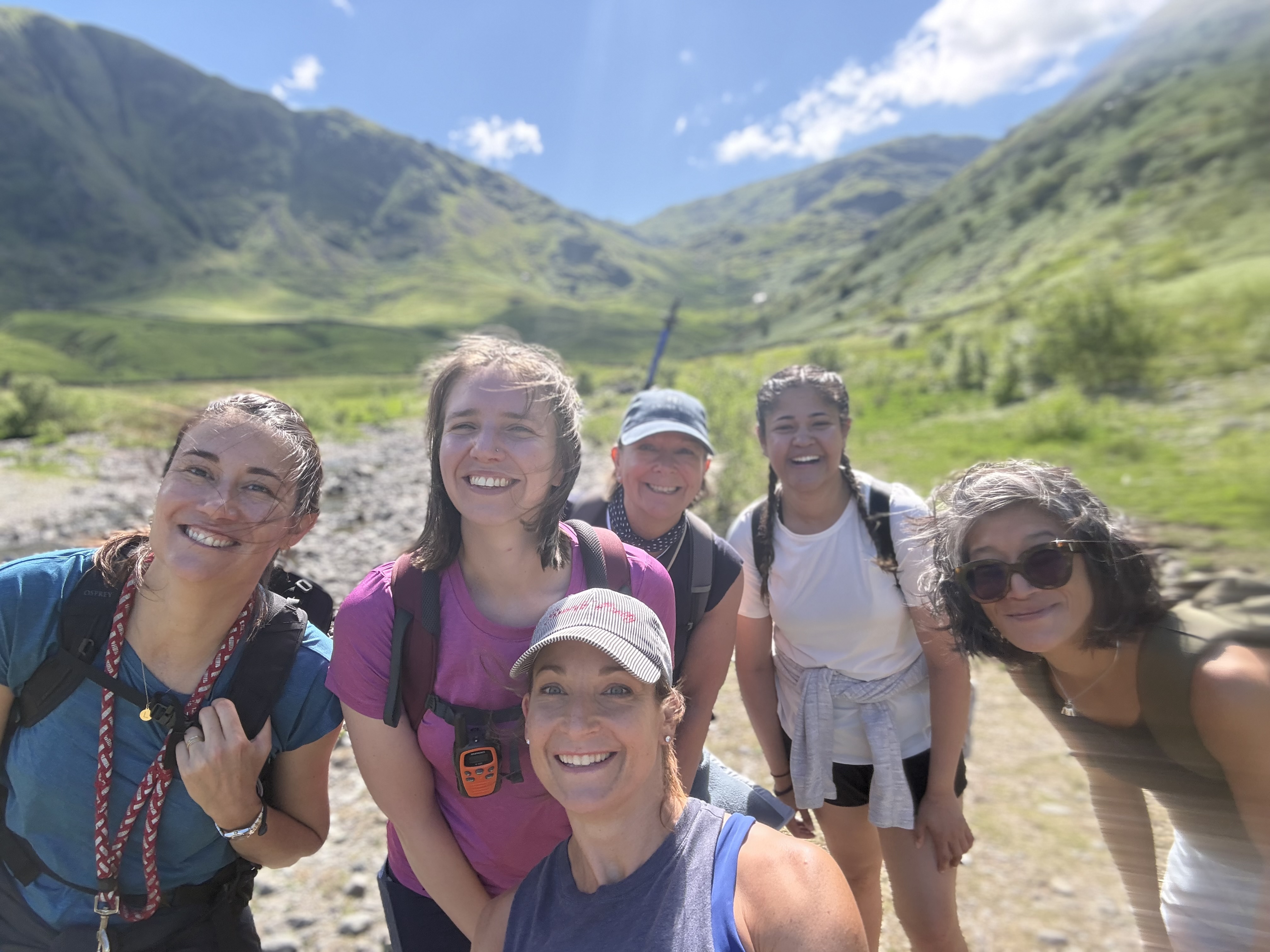 Joyful group selfie of six women mid-hike in a sun-drenched green valley