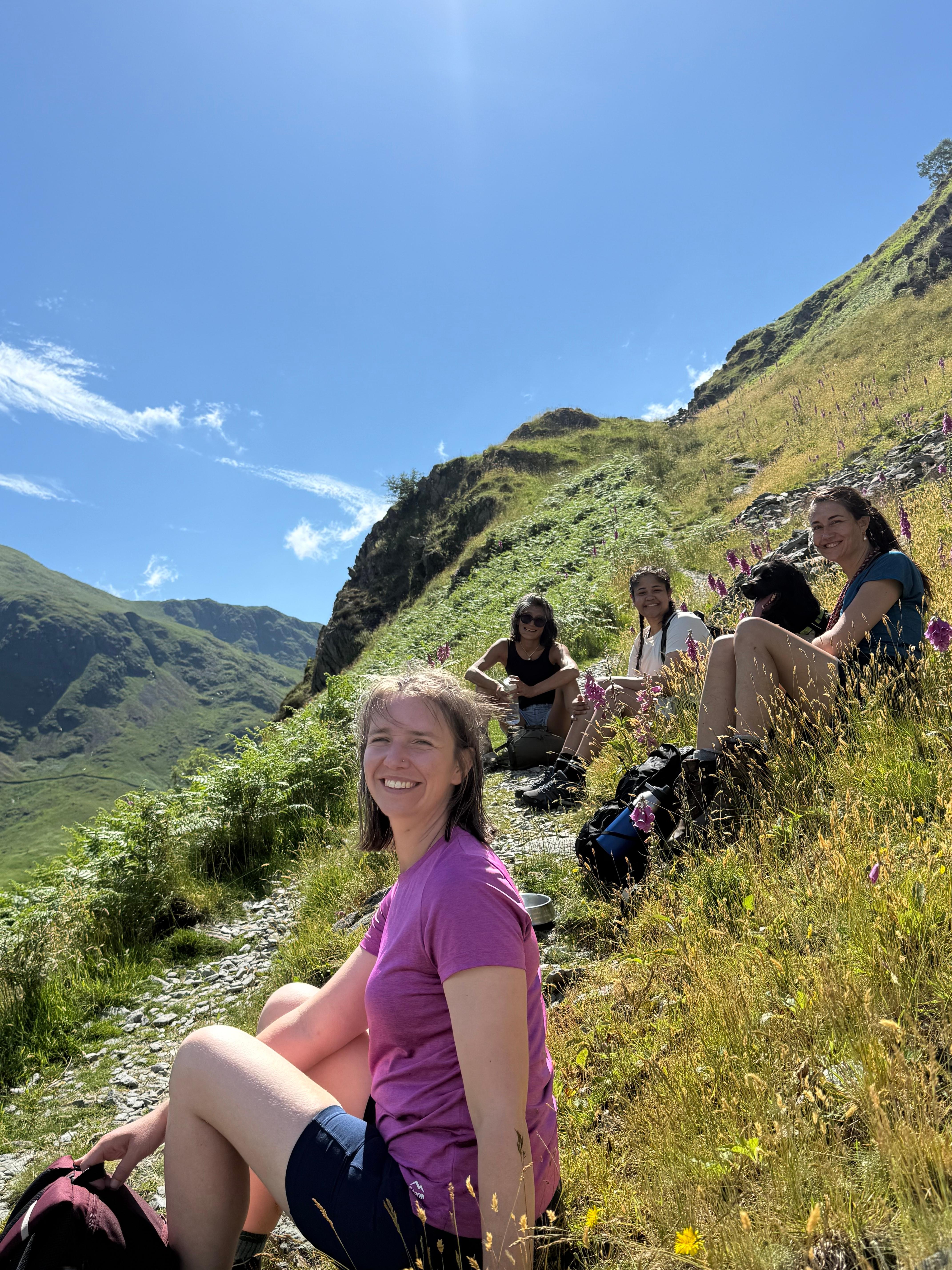 Women resting and smiling on a grassy hillside dotted with foxgloves during a sunny hike