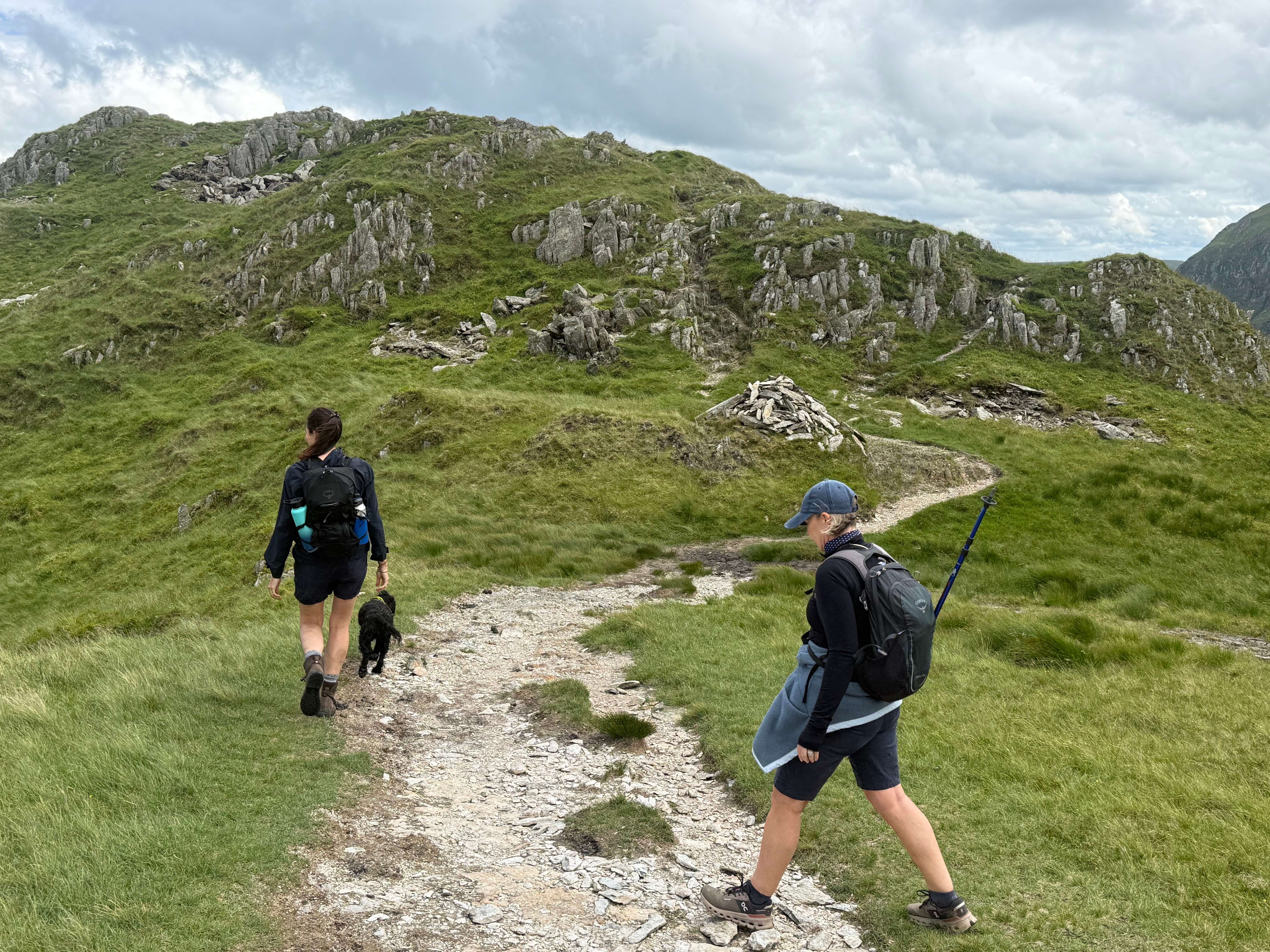 Two women and a black dog on a stony path heading toward a rocky green hill with a cairn