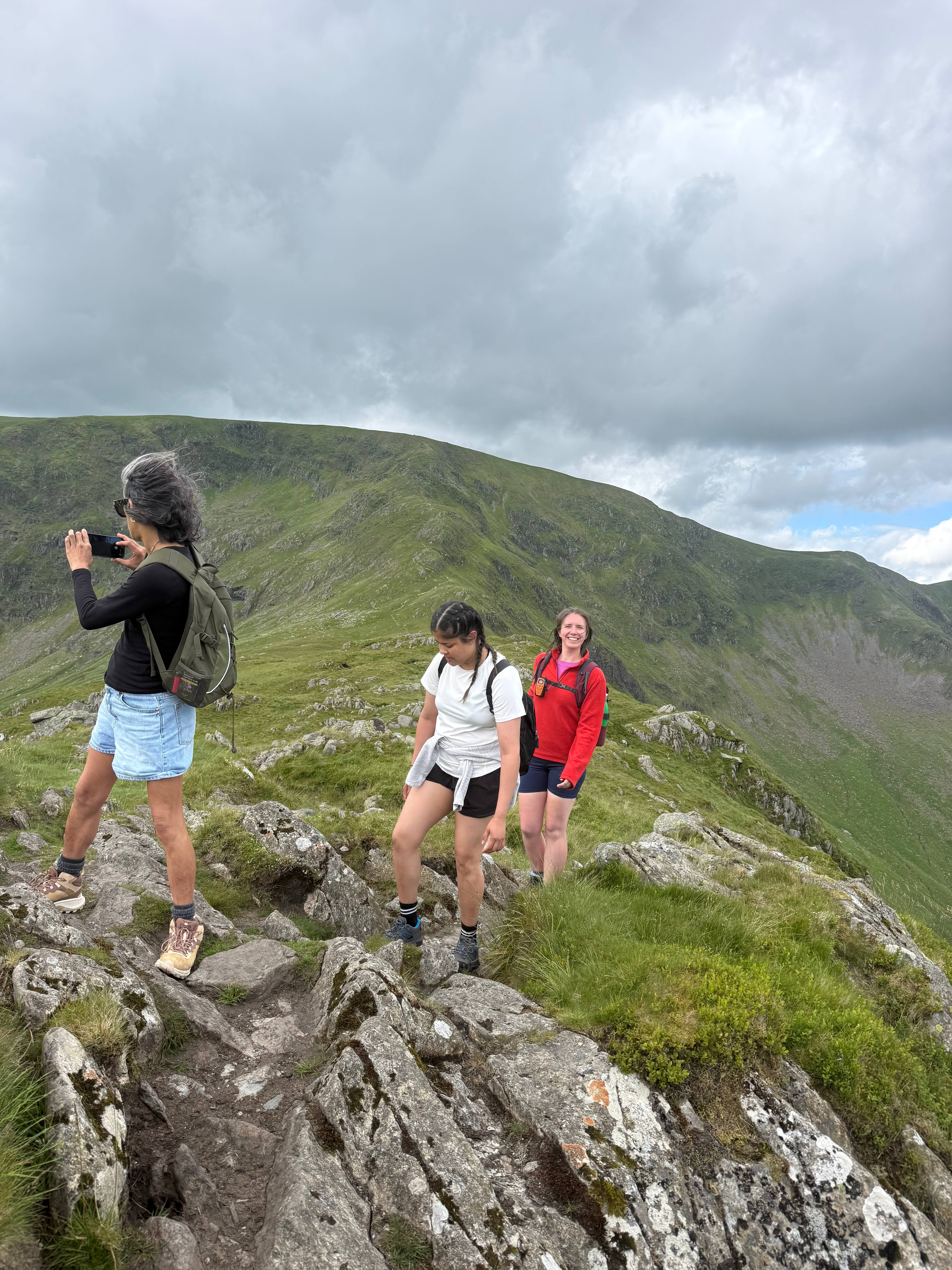 Three women on a rocky ridge, one photographing the sweeping green fells behind