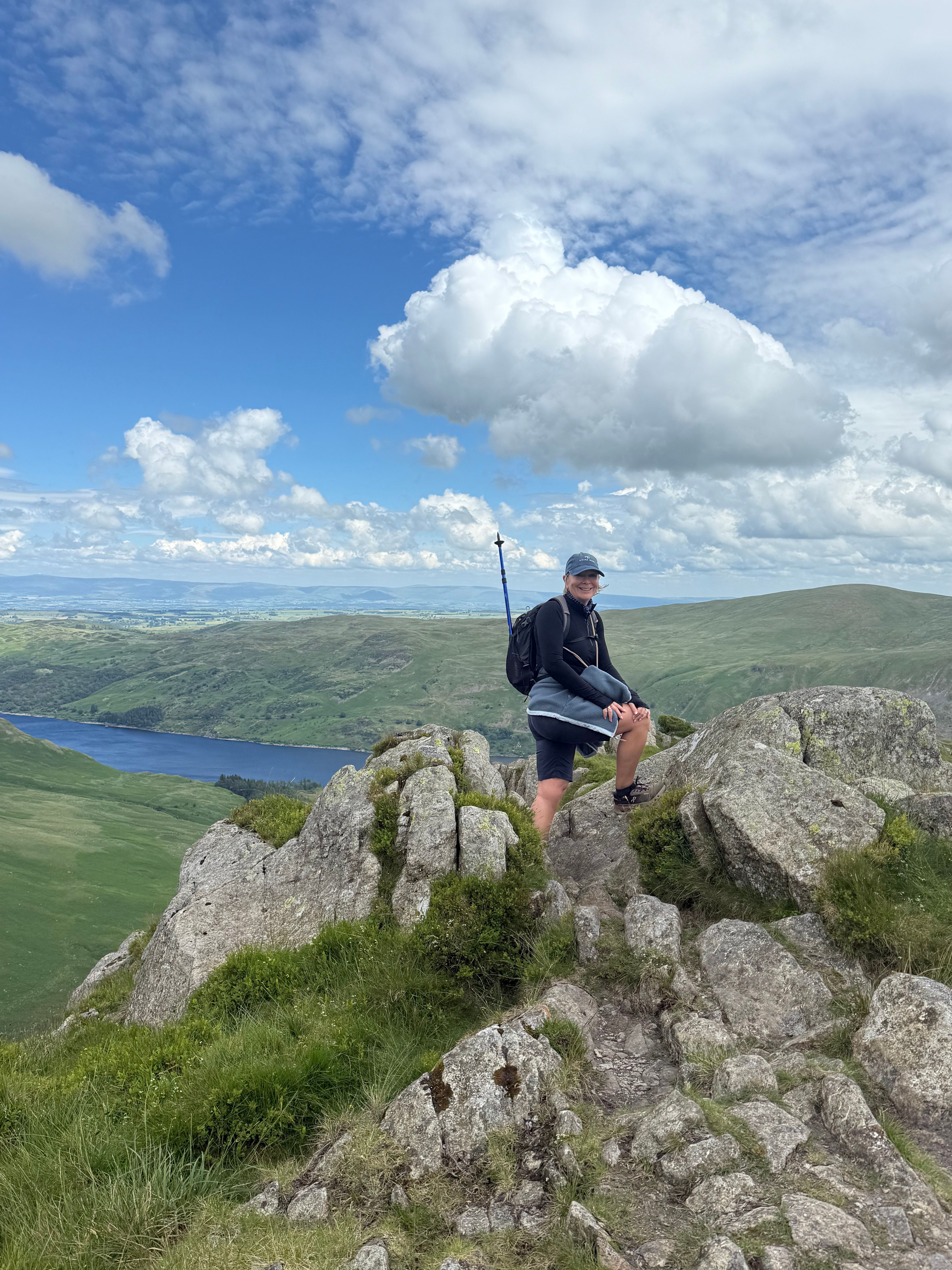 Smiling woman with a hiking pole and backpack standing on a rocky summit above Haweswater