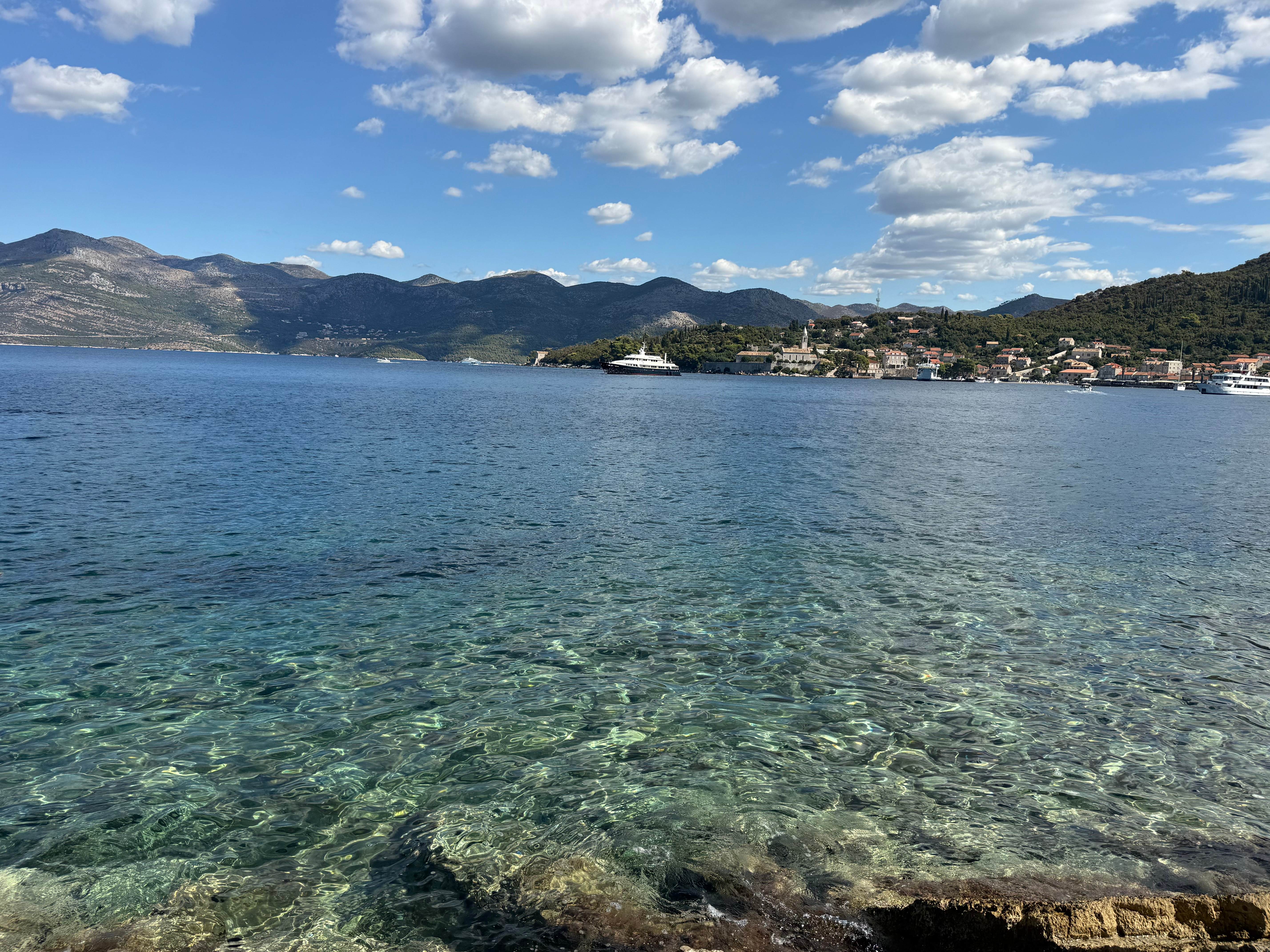 Clear turquoise water in the foreground looking across to a harbour village and mountains under white clouds