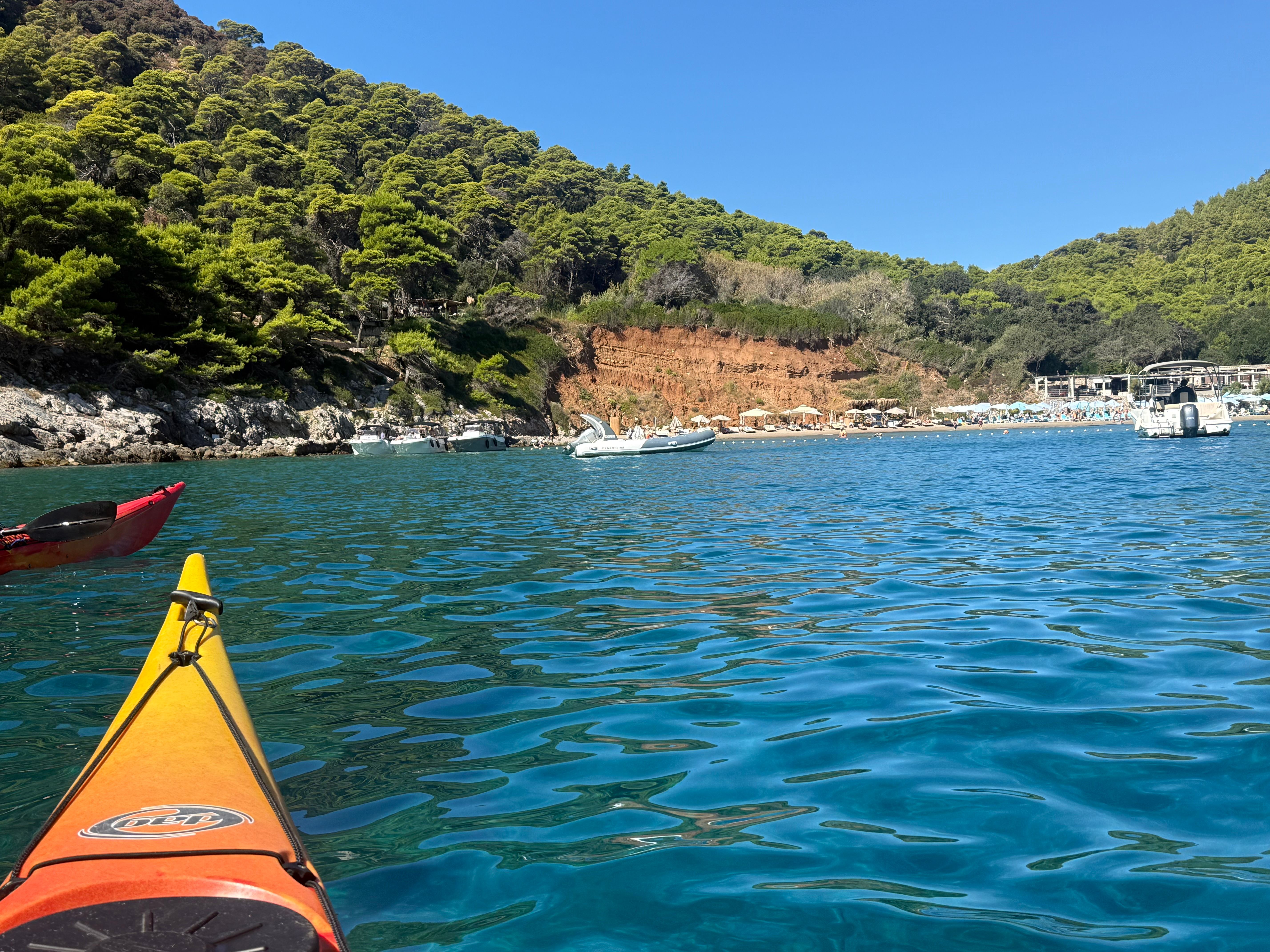 Kayak bow on blue water approaching a sandy beach beneath a red earth cliff and pine forest