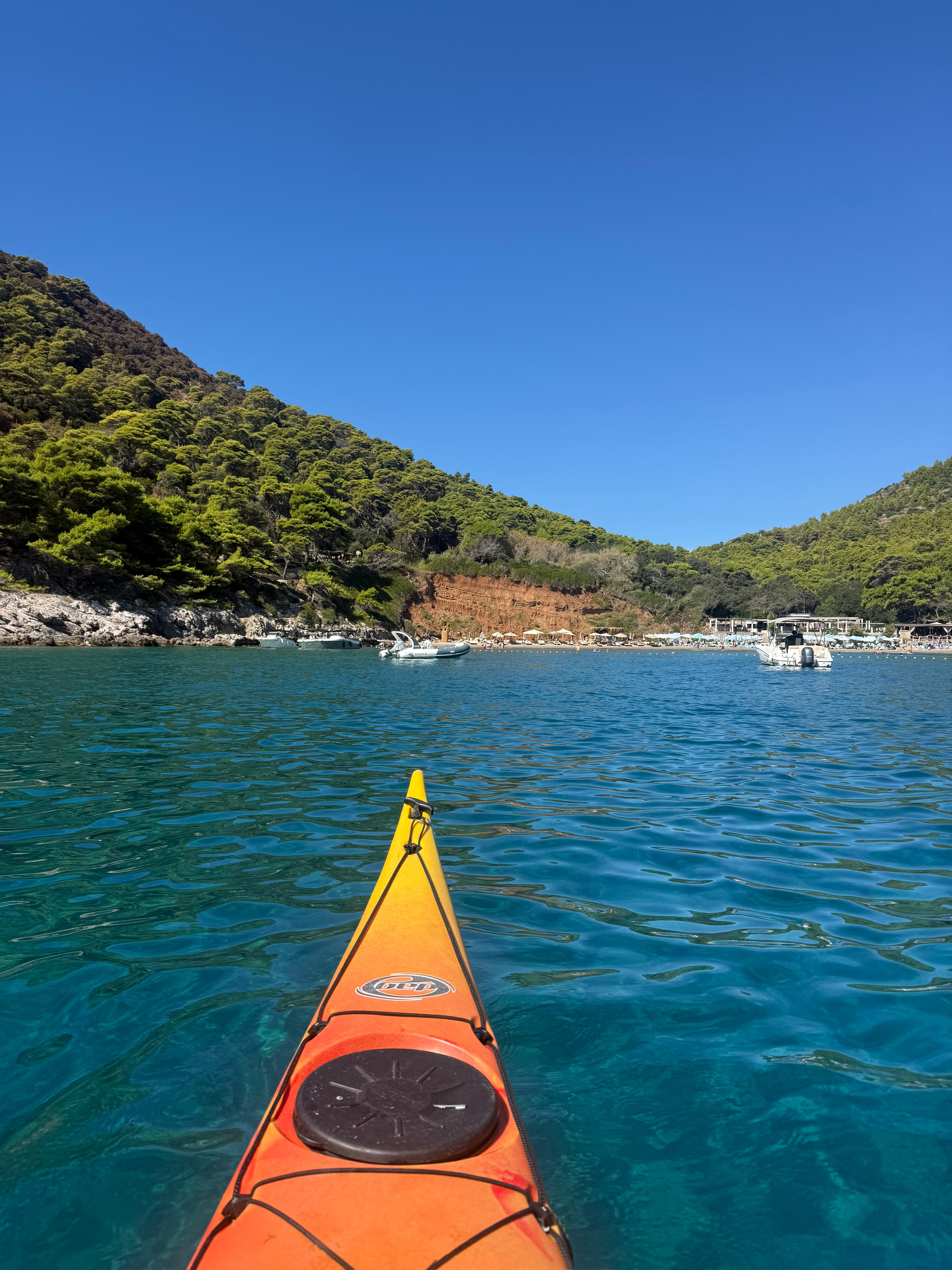 Bow of an orange sea kayak pointing towards a pine-covered cove with a sandy beach