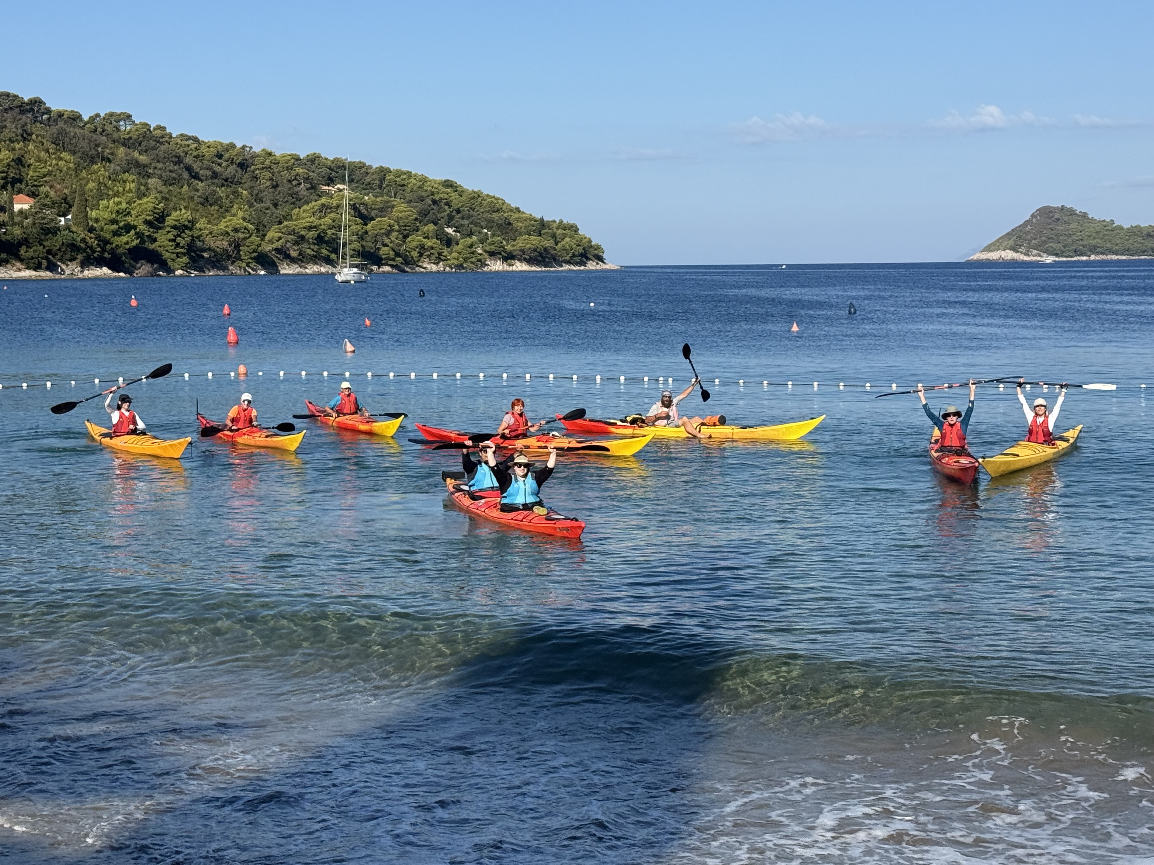 A group of women in red and yellow sea kayaks raising their paddles in a calm bay beneath a pine-covered headland