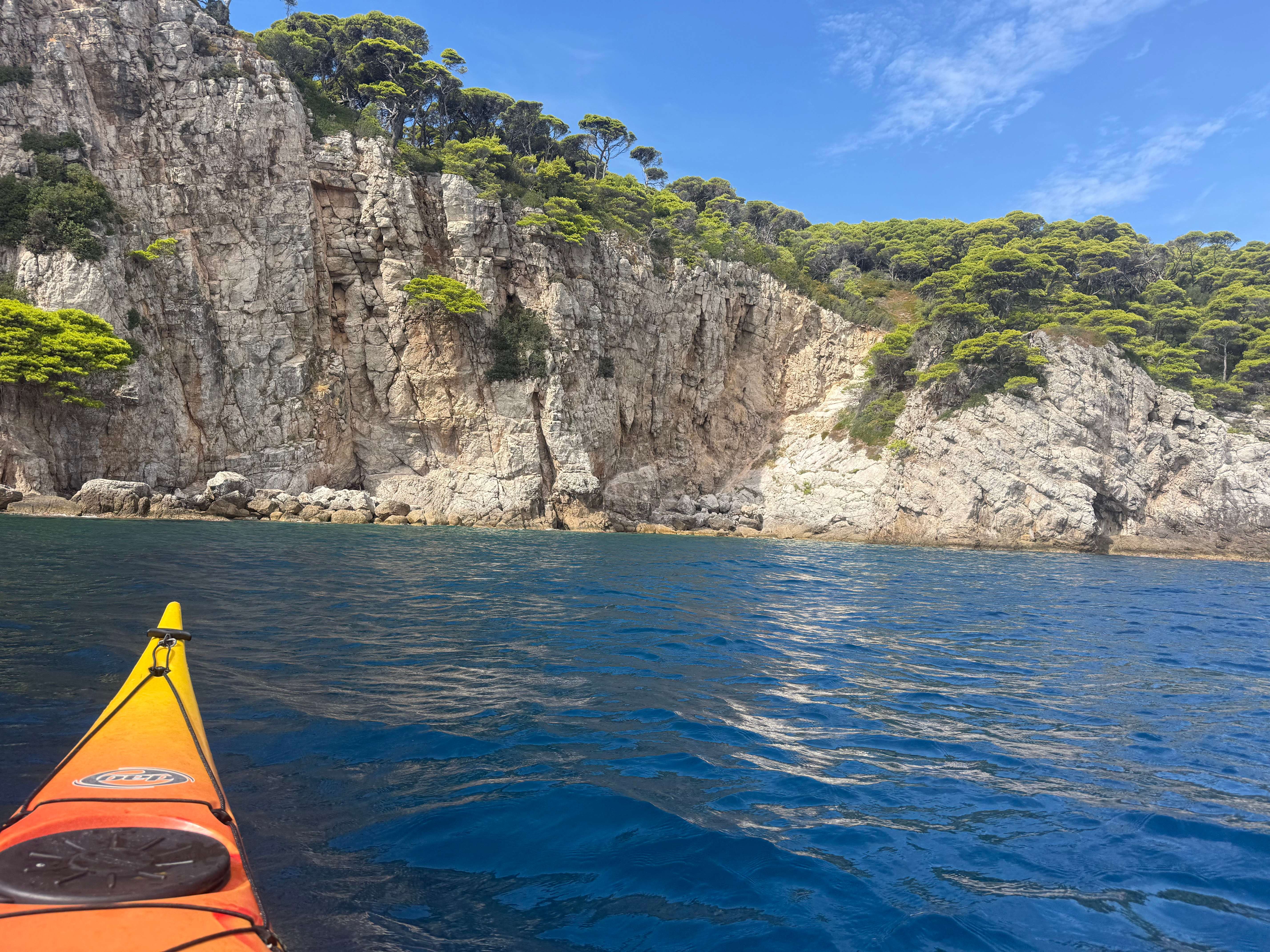 Bow of a yellow sea kayak approaching white limestone cliffs topped with pine trees