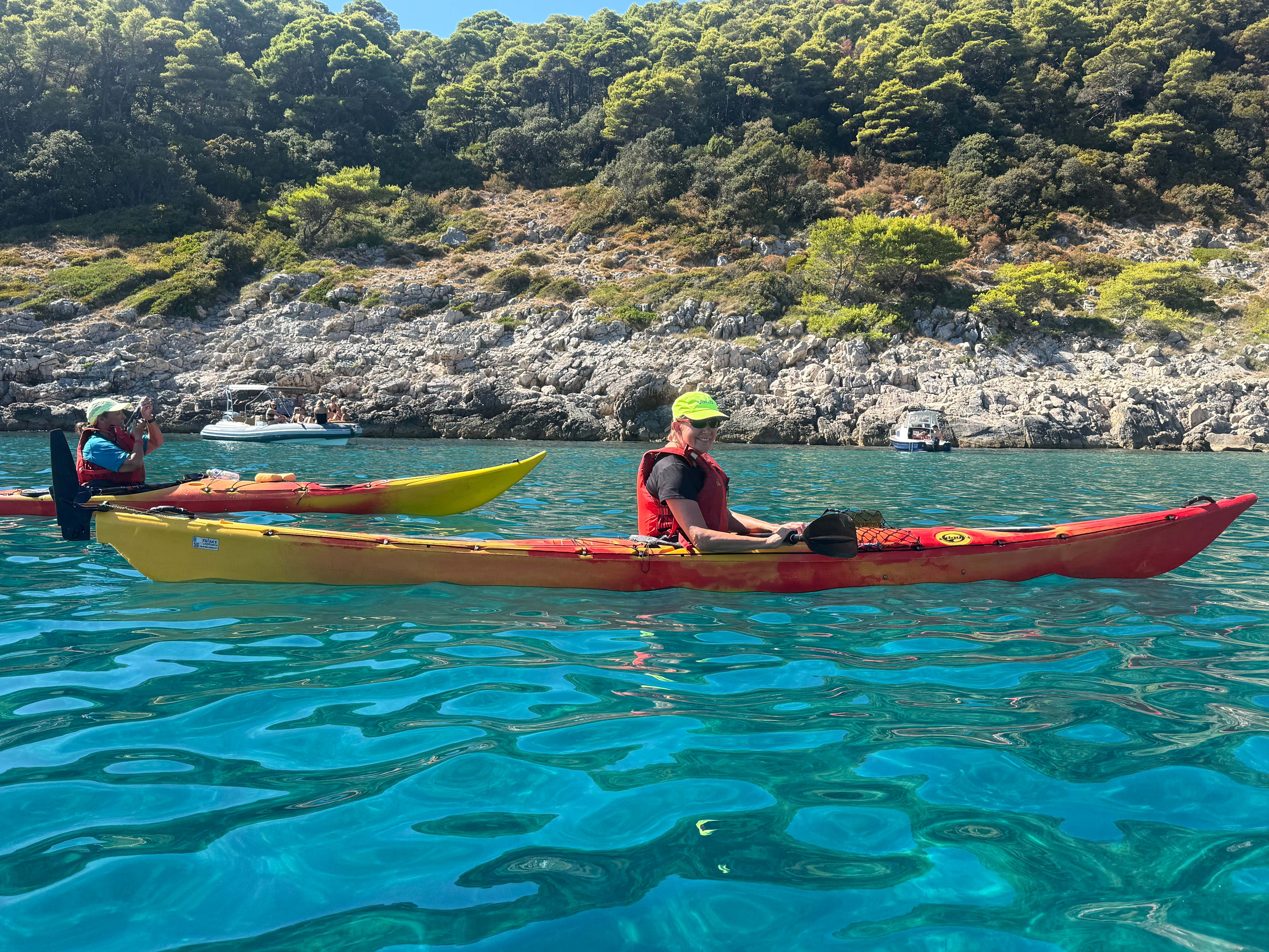 A smiling woman in a red and yellow sea kayak on turquoise water beneath a pine-covered limestone shore
