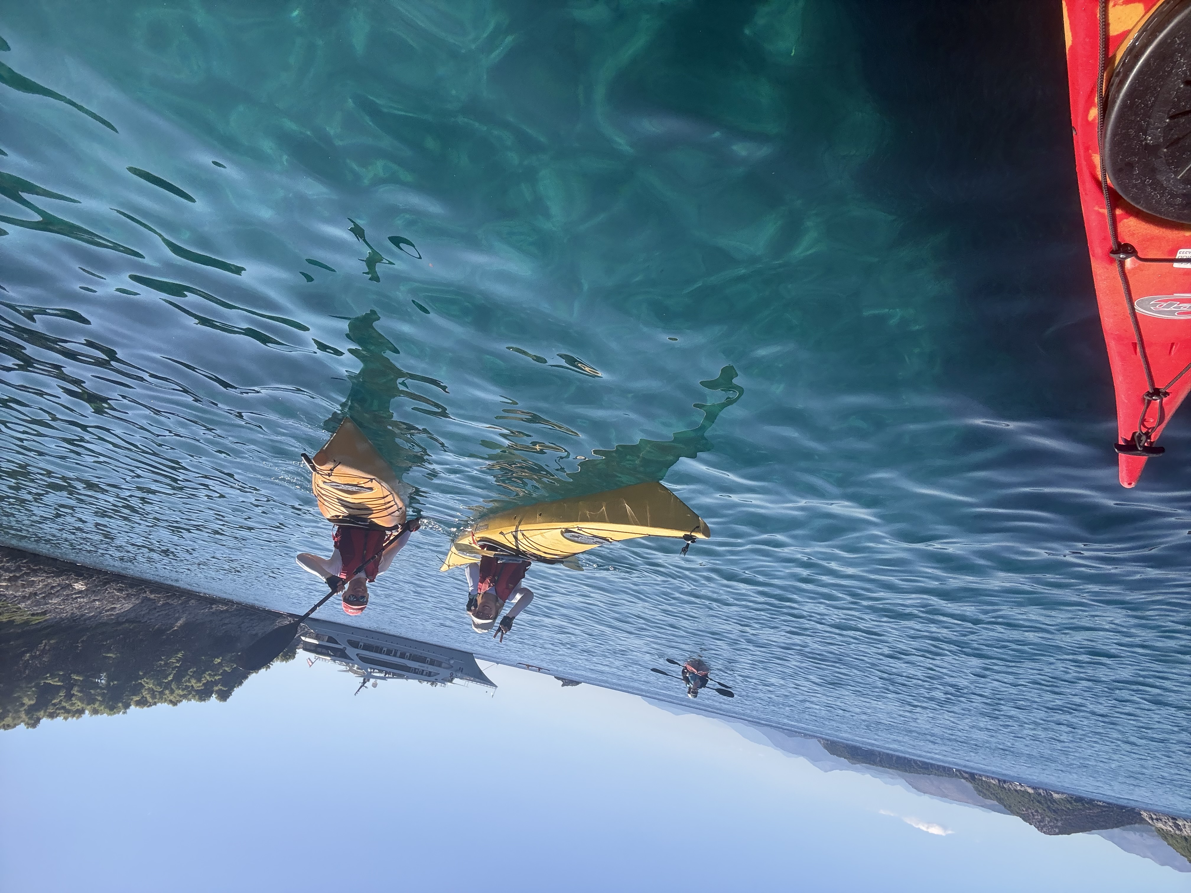 Two smiling kayakers in yellow kayaks on glass-clear turquoise water, a yacht anchored behind