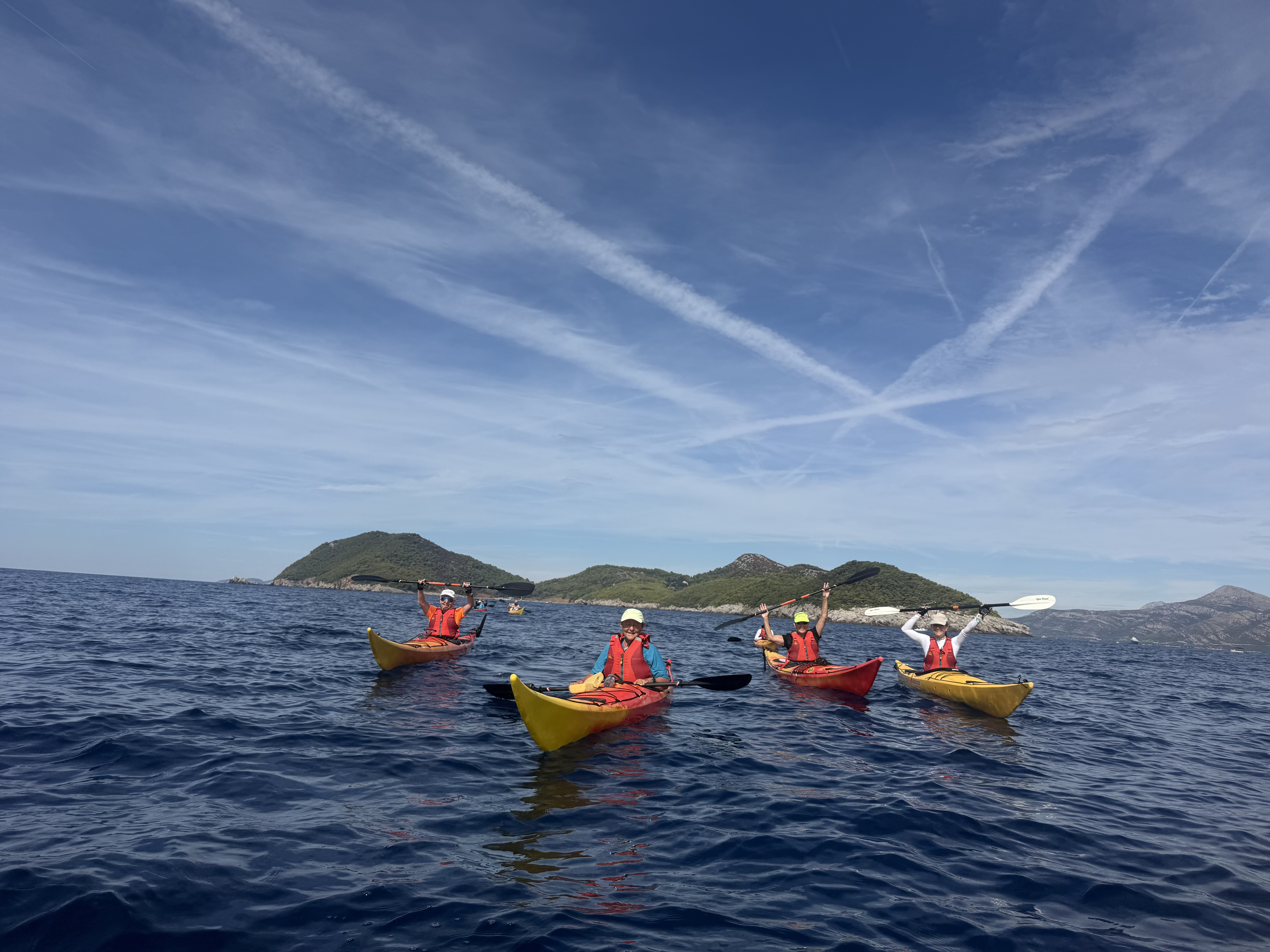 Four smiling kayakers holding paddles aloft on the open Adriatic with green islands behind