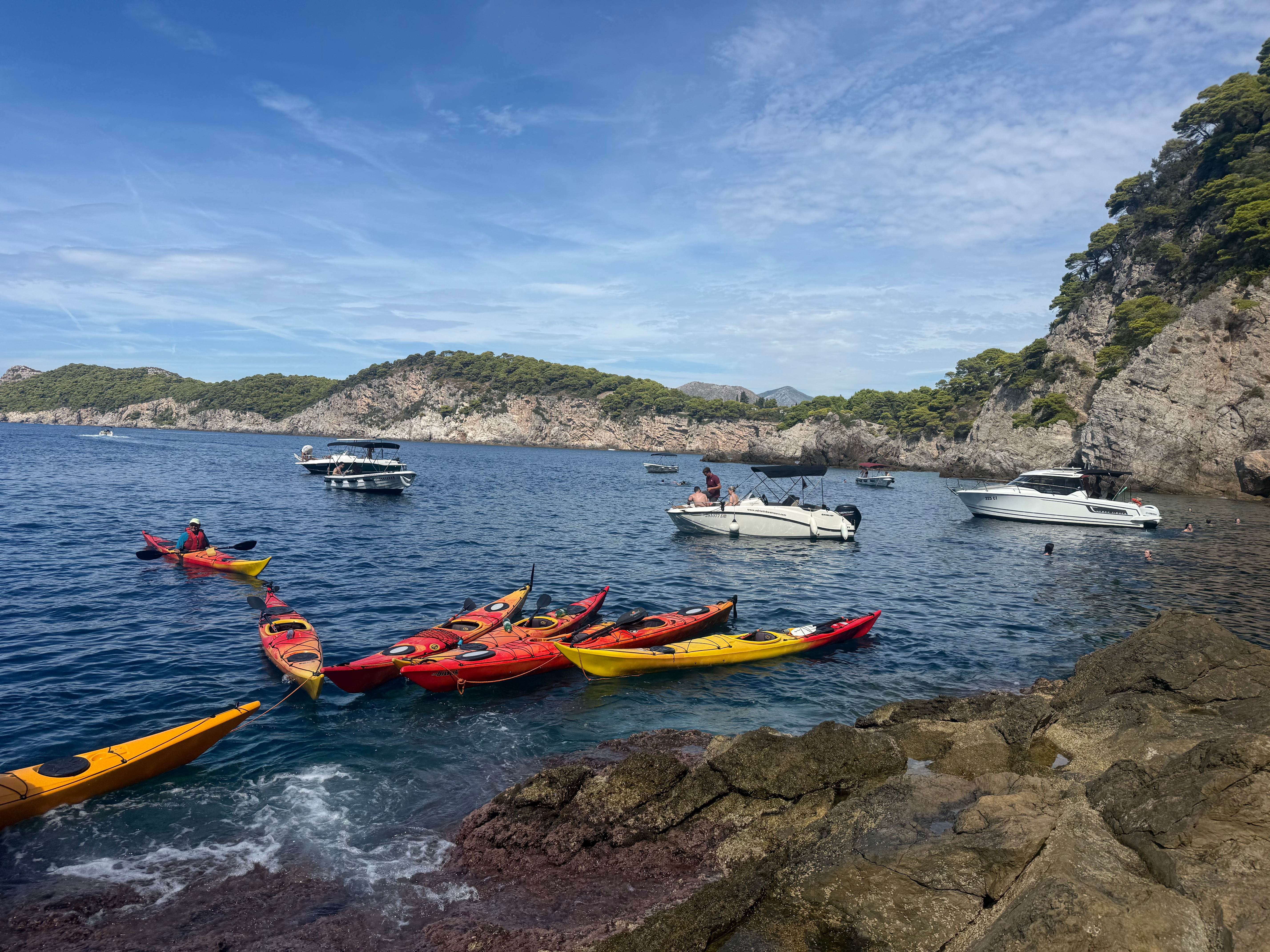 Colourful sea kayaks moored against rocks in a sheltered cove with boats and swimmers