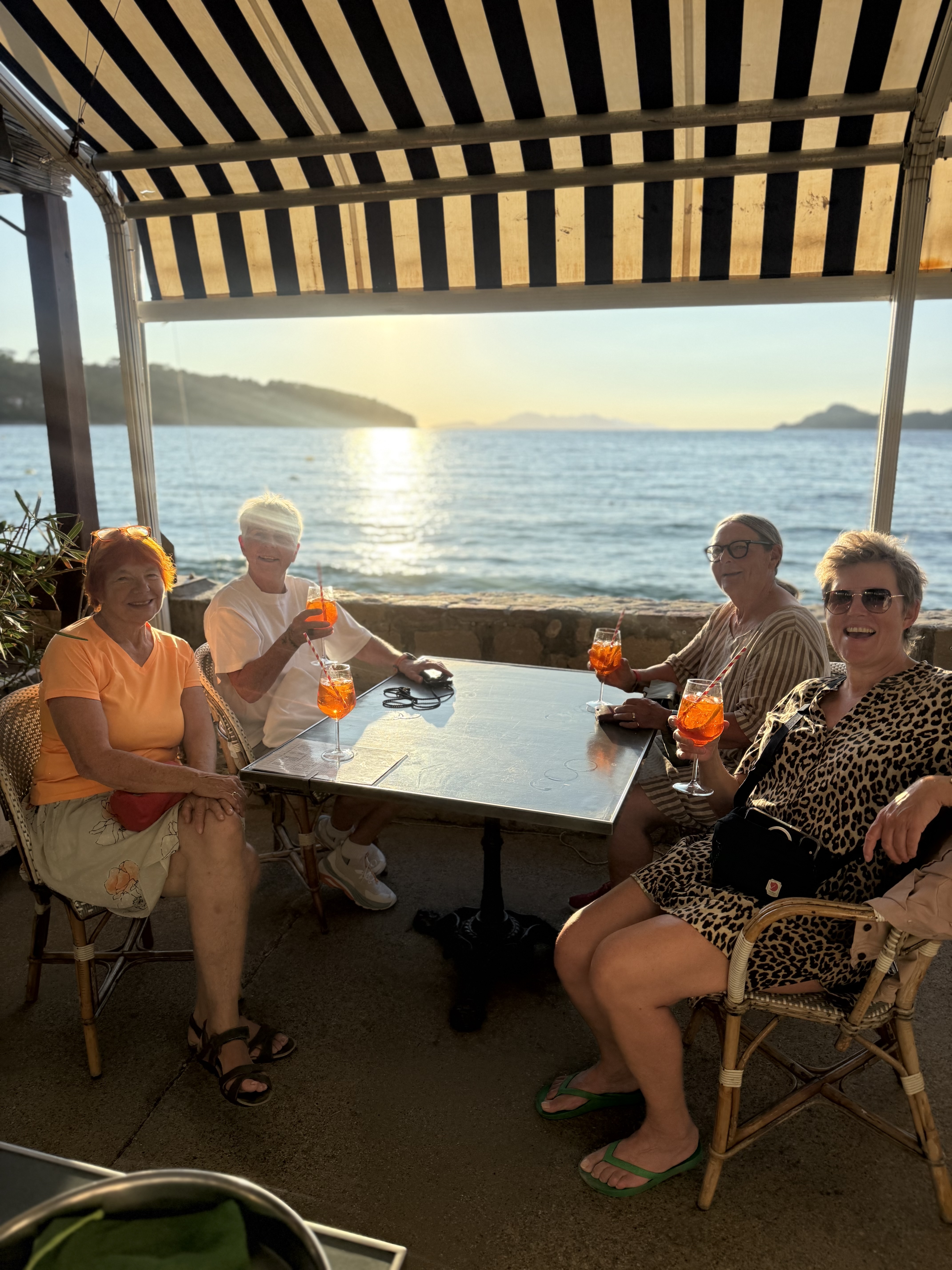 Four women raising spritz glasses at a seafront table under a striped awning at sunset